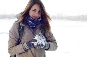 A young and joyful Caucasian girl in a brown coat holds a snowball in the background of a horizon line between the sky and a frozen lake in winter photo