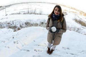 una joven y alegre chica caucásica con un abrigo marrón sostiene una bola de nieve frente a una línea de horizonte entre el cielo y un lago congelado en invierno. foto de ojo de pez