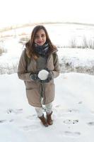 una joven y alegre chica caucásica con un abrigo marrón sostiene una bola de nieve frente a una línea de horizonte entre el cielo y un lago congelado en invierno. foto de ojo de pez