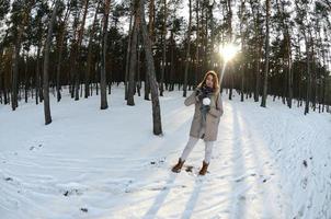 A young and joyful Caucasian girl in a brown coat holds a snowball in a snow-covered forest in winter. Fisheye Photo