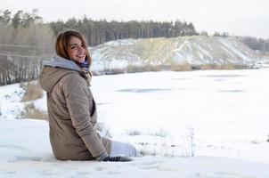 A young Caucasian girl in a brown coat is sitting near a cliff in the background of a horizon line between the sky and a frozen lake in winter time photo