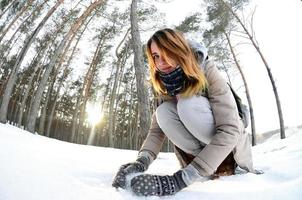 una joven y alegre chica caucásica con un abrigo marrón esculpe una bola de nieve en un bosque cubierto de nieve en invierno. juegos con nieve al aire libre. foto de ojo de pez