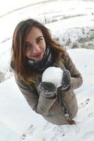 una joven y alegre chica caucásica con un abrigo marrón sostiene una bola de nieve frente a una línea de horizonte entre el cielo y un lago congelado en invierno. foto de ojo de pez