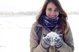 A young and joyful Caucasian girl in a brown coat holds a snowball in the background of a horizon line between the sky and a frozen lake in winter photo