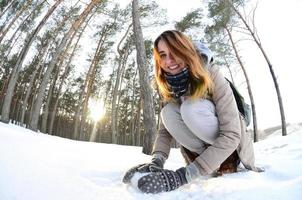 A young and joyful Caucasian girl in a brown coat sculpts a snowball in a snow-covered forest in winter. Games with snow in the open air. Fisheye Photo