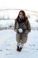 una joven y alegre chica caucásica con un abrigo marrón sostiene una bola de nieve frente a una línea de horizonte entre el cielo y un lago congelado en invierno. foto de ojo de pez