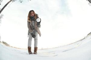 una joven y alegre chica caucásica con un abrigo marrón sostiene una bola de nieve frente a una línea de horizonte entre el cielo y un lago congelado en invierno. foto de ojo de pez