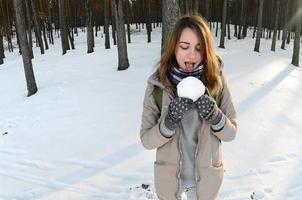 una joven y alegre chica caucásica con un abrigo marrón sostiene una bola de nieve en un bosque cubierto de nieve en invierno. foto de ojo de pez