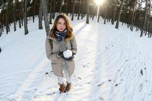 A young and joyful Caucasian girl in a brown coat holds a snowball in a snow-covered forest in winter. Fisheye Photo