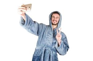 El retrato de un joven caucásico con barba y albornoz azul muestra un gesto de victoria y hace un selfie aislado de fondo blanco. foto