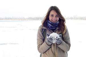 A young and joyful Caucasian girl in a brown coat holds a snowball in the background of a horizon line between the sky and a frozen lake in winter photo