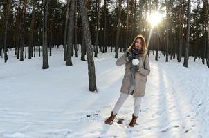 A young and joyful Caucasian girl in a brown coat holds a snowball in a snow-covered forest in winter. Fisheye Photo