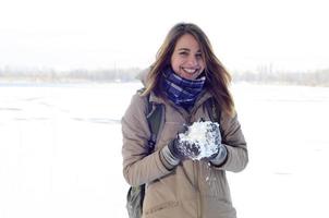 A young and joyful Caucasian girl in a brown coat holds a snowball in the background of a horizon line between the sky and a frozen lake in winter photo