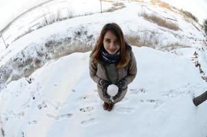 una joven y alegre chica caucásica con un abrigo marrón sostiene una bola de nieve frente a una línea de horizonte entre el cielo y un lago congelado en invierno. foto de ojo de pez