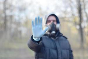Blurred portrait of young man in protective gas mask and rubber disposable gloves shows stop gesture outdoors in spring wood photo