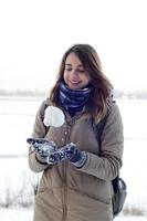 A young and joyful Caucasian girl in a brown coat holds a snowball in the background of a horizon line between the sky and a frozen lake in winter photo