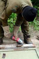 Rectificado eléctrico de ruedas en estructura de acero al aire libre. viejo trabajador manual con guantes protectores cortando puertas de metal con molinillo foto