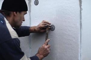 un trabajador anciano obstruye una espiga en un paraguas de plástico montado en una pared de espuma de poliestireno. el proceso de fijación de placas de poliestireno expandido. calentamiento de la fachada del edificio foto