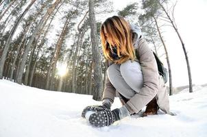 A young and joyful Caucasian girl in a brown coat sculpts a snowball in a snow-covered forest in winter. Games with snow in the open air. Fisheye Photo
