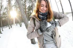 A young and joyful Caucasian girl in a brown coat sculpts a snowball in a snow-covered forest in winter. Games with snow in the open air. Fisheye Photo