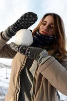 A young and joyful Caucasian girl in a brown coat holds a snowball in front of a horizon line between the sky and a frozen lake in winter. Fisheye Photo