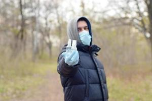 joven con máscara protectora muestra botellas de spray desinfectante al aire libre en madera de primavera foto