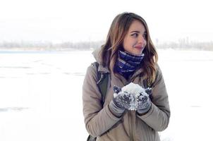 A young and joyful Caucasian girl in a brown coat holds a snowball in the background of a horizon line between the sky and a frozen lake in winter photo