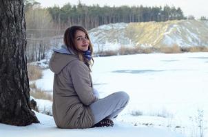 A young Caucasian girl in a brown coat is sitting near a cliff in the background of a horizon line between the sky and a frozen lake in winter time photo