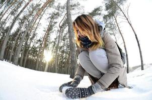 A young and joyful Caucasian girl in a brown coat sculpts a snowball in a snow-covered forest in winter. Games with snow in the open air. Fisheye Photo