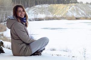 A young Caucasian girl in a brown coat is sitting near a cliff in the background of a horizon line between the sky and a frozen lake in winter time photo