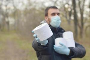 Covidiot concept. Young man in protective mask holds many rolls of toilet paper outdoors in spring wood photo