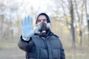 Blurred portrait of young man in protective gas mask and rubber disposable gloves shows stop gesture outdoors in spring wood photo