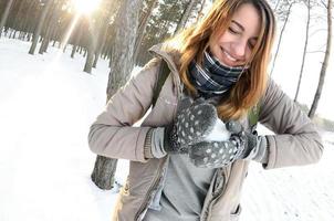 A young and joyful Caucasian girl in a brown coat sculpts a snowball in a snow-covered forest in winter. Games with snow in the open air. Fisheye Photo