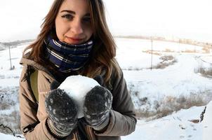 A young and joyful Caucasian girl in a brown coat holds a snowball in front of a horizon line between the sky and a frozen lake in winter. Fisheye Photo