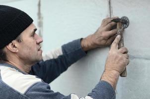 An elderly workman clogs a dowel into a plastic umbrella mount in a styrofoam wall. The process of fixing expanded polystyrene plates. Warming of the facade of the building photo