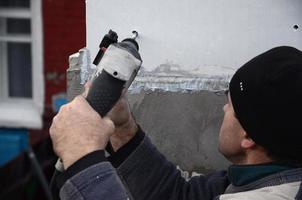 An elderly workman drills a hole in a styrofoam wall for the subsequent installation of a plastic reinforcing dowel. Creating holes in the wall with a drill. Warming of the building facade photo