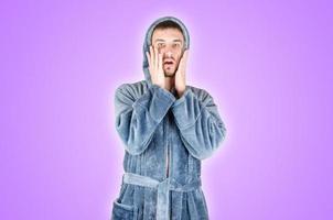 Portrait of young caucasian bearded man in blue bathrobe be scared isolated on violet background photo