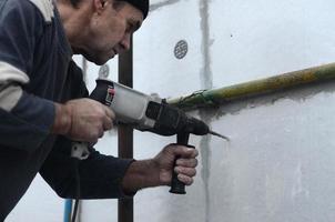 un trabajador anciano perfora un agujero en una pared de espuma de poliestireno para la posterior instalación de una espiga de refuerzo de plástico. creando agujeros en la pared con un taladro. calentamiento de la fachada del edificio foto