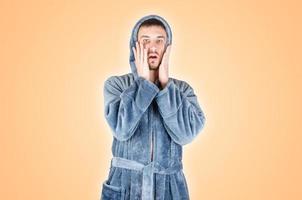 Portrait of young caucasian bearded man in blue bathrobe be scared isolated on orange background photo