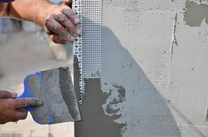 Hands of an old manual worker with wall plastering tools renovating house. Plasterer renovating outdoor walls and corners with spatula and plaster. Wall insulation. Construction finishing works photo