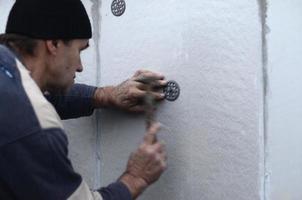 un trabajador anciano obstruye una espiga en un paraguas de plástico montado en una pared de espuma de poliestireno. el proceso de fijación de placas de poliestireno expandido. calentamiento de la fachada del edificio foto