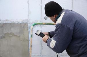 un trabajador anciano perfora un agujero en una pared de espuma de poliestireno para la posterior instalación de una espiga de refuerzo de plástico. creando agujeros en la pared con un taladro. calentamiento de la fachada del edificio foto