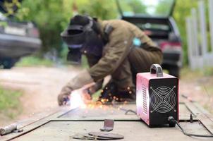 Old man welder in brown uniform, welding mask and welders leathers, weld metal door with arc welding machine outdoors photo