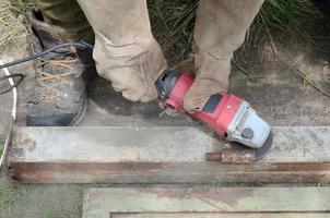 Rectificado eléctrico de ruedas en estructura de acero al aire libre. viejo trabajador manual con guantes protectores cortando puertas de metal con molinillo foto