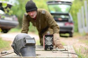 Old man welder in brown uniform prepares metal door surface for welding with arc welding machine outdoors photo