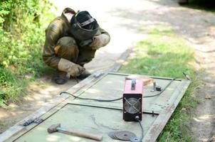 Old man welder in brown uniform, welding mask and welders leathers, weld metal door with arc welding machine outdoors photo