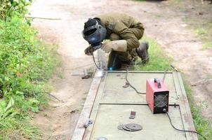 Old man welder in brown uniform, welding mask and welders leathers, weld metal door with arc welding machine outdoors photo