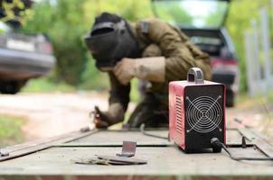 Old man welder in brown uniform, welding mask and welders leathers, weld metal door with arc welding machine outdoors photo