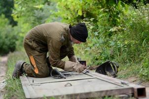 Old man welder in brown uniform prepares metal door surface for welding with arc welding machine outdoors photo