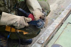 Rectificado eléctrico de ruedas en estructura de acero al aire libre. viejo trabajador manual con guantes protectores cortando puertas de metal con molinillo foto